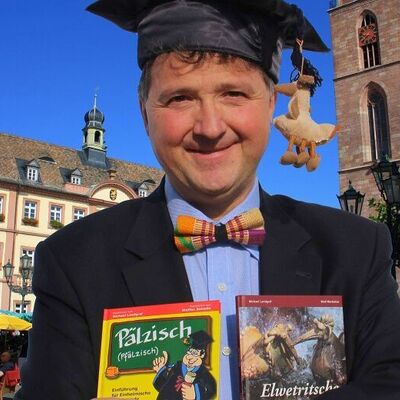 Bild vergr&ouml;&szlig;ern: Mann mit einem Gelehrtenhut hält zwei Bücher in den Händen und lächelt in die Kamera auf dem Marktplatz