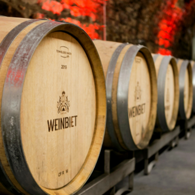 Eine Reihe von Barriquefässern mit dem Logo der WEINBIET-Manufaktur in einem rot illuminierten Gewölbekeller. 