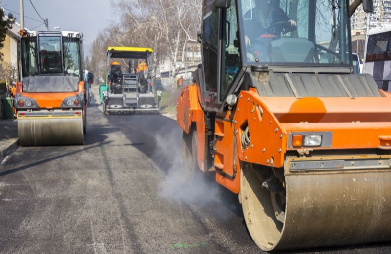 Im Vordergrund eine Walze, die frischen Asphalt plättet. Im Hintergrund weitere Baustellenfahrzeuge.