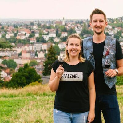 Bild vergr&ouml;&szlig;ern: Ein junges Paar auf einer Wiese mit gefüllten Weingläsern in der Hand und im Hintergrund Blick auf eine Stadt - Haardt Core Pfalz
