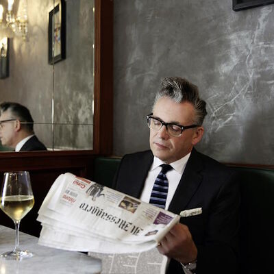 Der Musiker Götz Alsmann sitz in einem Restaurant und liest eine Zeitung. Vor ihm steht ein Glas gefüllt mit Weißwein.
