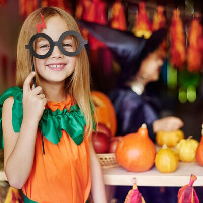 Ein Mädchen in einem Kürbiskostüm schaut in die Kamera und hält sich eine schwarze Brille mit einem roten Blitz darauf vor die Augen. Im Hintergrund sind weitere verkleidete Kinder und Kürbisse zu sehen.