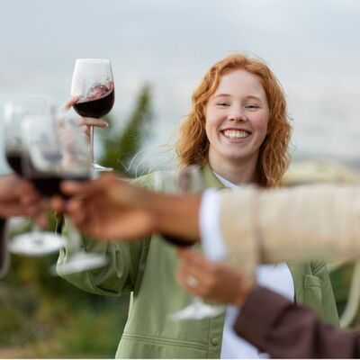 Eine lachende Frau hält ein Glas Rotwein in der Hand und prostet andren Menschen zu.