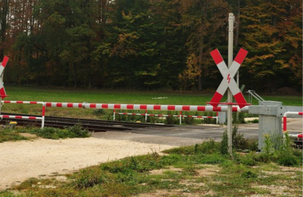 Das Bild zeigt einen geschlossenen Bahnübergang auf einer Landstraße. Im Hintergrund ist ein Waldgebiet zu sehen.
