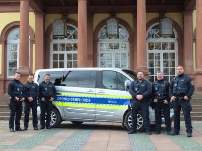 Bild vergr&ouml;&szlig;ern: Drei Mitarbeiter des kommunalen Vollzugsdienstes stehen in Reihe aufgestellt neben einem Fahrzeug der Ordnungsbehörde Neustadt an der Weinstraße. Das Fahrzeug ist vor dem Neustadter Saalbau geparkt.