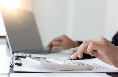 Bild vergr&ouml;&szlig;ern: Nahaufnahme von einer Person, die am Schreibtisch arbeitet und einen Stift hält, vor sich einen Laptop