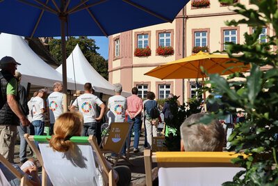 Bild vergr&ouml;&szlig;ern: Personen auf Liegestühlen wurden von hinten fotografiert. Im Hintergrund befindet sich rechts ein Ausschnitt des Rathauses sowie weitere Besuchende auf dem Marktplatz.