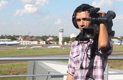 Bild vergr&ouml;&szlig;ern: Ein Kameramann steht mit seiner Ausrüstung vor einem Flughafen