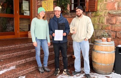 Bild vergr&ouml;&szlig;ern: Eine Mitarbeiterin der TI und zwei Betreiber des Weinbiethauses sind im Winter vor der "Woibiet"-Hütte zu sehen und halten das Schild und die Zertifizierungsurkunde in den Händen.