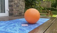 Ein orangefarbener Ball liegt auf einer blauen Yogamatte auf einer Holzterrasse, im Hintergrund ist ein Garten zu sehen.