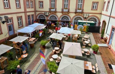 Bild vergr&ouml;&szlig;ern: Blick in den Neustadter Rathausinnenhof. Hier sind Tische und Stühle gestellt, es gibt einen Ausschank. Fahnen und Banner weisen auf "1832. Das Fest der Demokratie" hin. Das Bild verlinkt zu weiteren Informationen über das Weindorf im Rathausinnenhof und die Town Hall Bühne (Treffpunktformat) während des Demokratiefestes am 30.05.2026