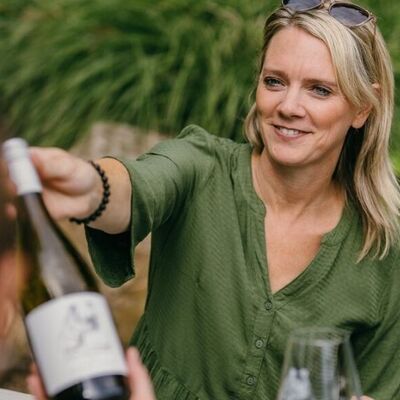 Eine blonde Frau in einem grünen Shirt reicht eine Weinflasche über den Tisch.