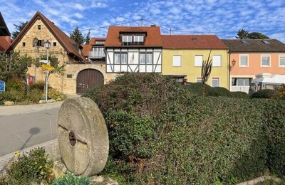 Historische Loblocher Gebäudefront im Weindorf Gimmeldingen. Im Vordergrund liegt ein Mühlstein der ehemaligen Loblocher Mühle, umgeben von grünen Sträuchern.