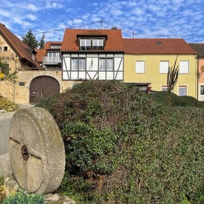 Historische Loblocher Gebäudefront im Weindorf Gimmeldingen. Im Vordergrund liegt ein Mühlstein der ehemaligen Loblocher Mühle, umgeben von grünen Sträuchern.