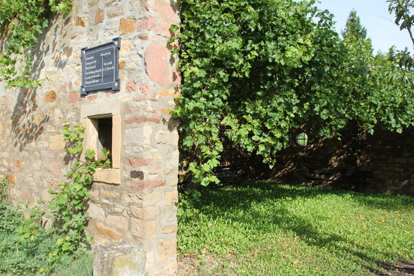 Bild vergr&ouml;&szlig;ern: Alte Mauer im Grünen mit kleinem Sandsteinfenster. Darüber altes Ortseingangsschild.